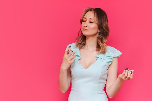 Photo a young caucasian pretty brownhaired woman with her eyes closed in a light blue stylish dress holding perfume bottle in her hand and applying it on her neck isolated on a bright color pink wall