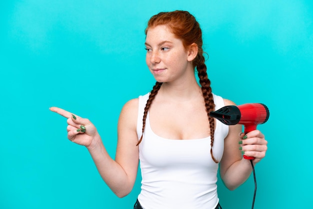 Young caucasian reddish holding a hairdryer isolated on blue background pointing to the side to present a product