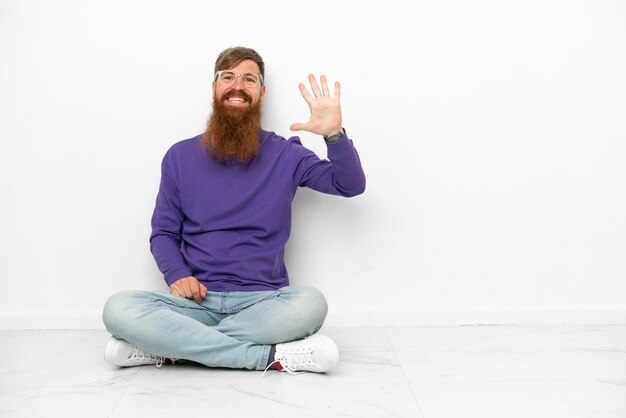 Young caucasian reddish man sitting on the floor isolated on white background counting five with fingers