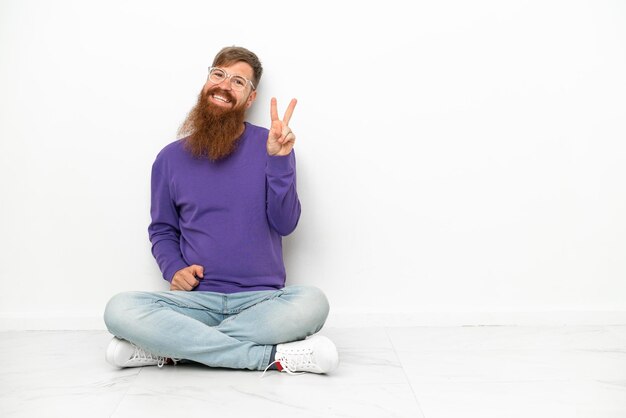 Young caucasian reddish man sitting on the floor isolated on white background smiling and showing victory sign