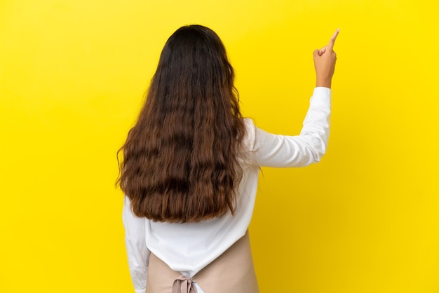 Young caucasian restaurant waiter isolated on yellow background pointing back with the index finger