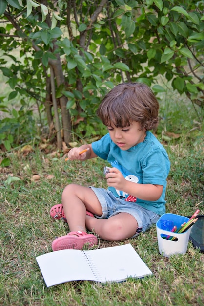 Young Caucasian school boy sitting in park outdoor doing school homework Child kid writing in notebook with pencil outside Self education learning studying Early development for children