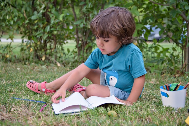 Young Caucasian school boy sitting in park outdoor doing school homework Child kid writing in notebook with pencil outside Self education learning studying Early development for children