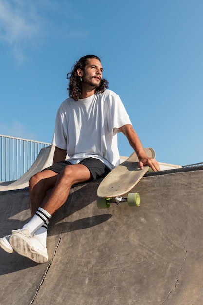 Young caucasian skater with long black hair sitting at the top of the skate ramp