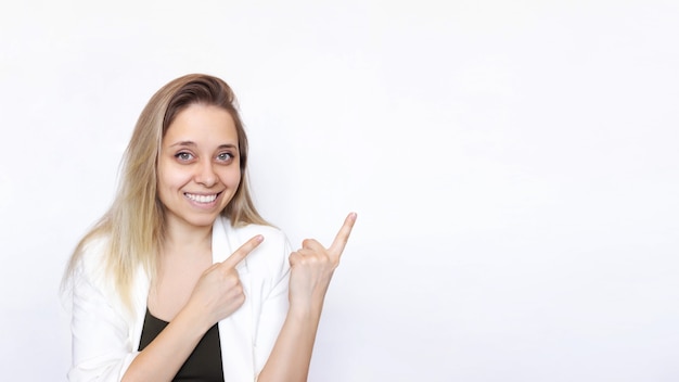 A young caucasian smiling blonde woman in a white jacket points at copy space on white background