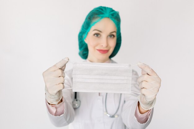 Young Caucasian smiling woman doctor in cap wearing protection face mask against infections, bacteria and virus, isolated on white. Focus on the mask in hands. Coronavirus covid-19 concept.