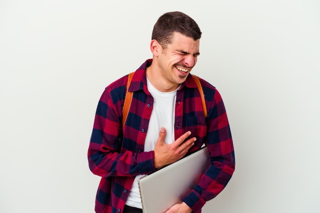 Young caucasian student man holding a laptop isolated on white background laughs happily and has fun keeping hands on stomach.