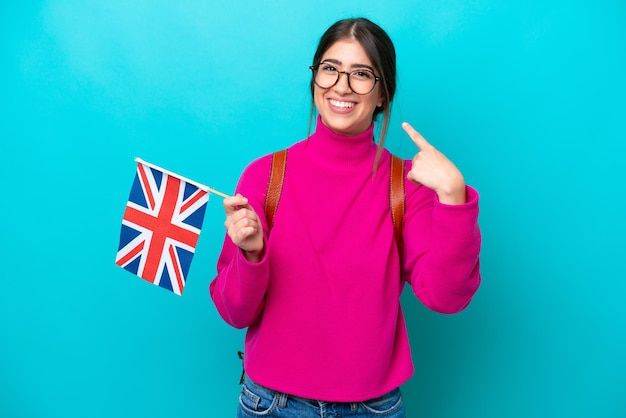 Young caucasian student woman holding English flag isolated on blue background giving a thumbs up gesture