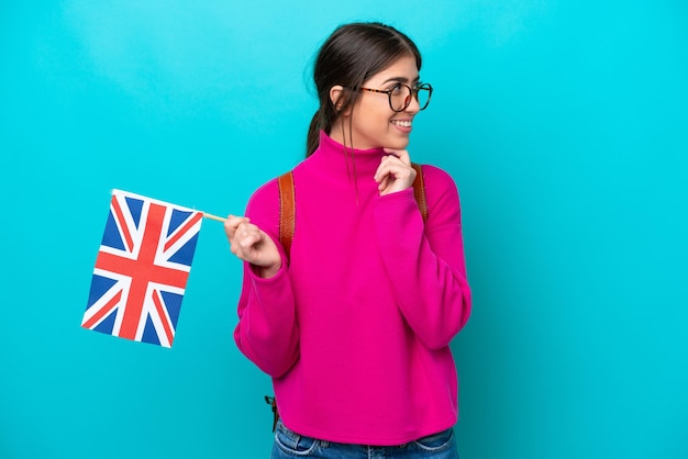 Young caucasian student woman holding English flag isolated on blue background looking to the side and smiling