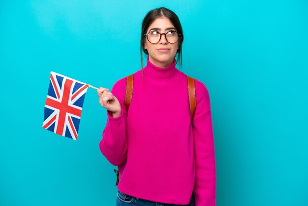 Young caucasian student woman holding English flag isolated on blue background and looking up