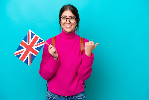 Young caucasian student woman holding English flag isolated on blue background pointing to the side to present a product