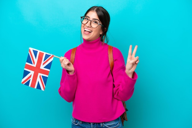 Young caucasian student woman holding English flag isolated on blue background smiling and showing victory sign