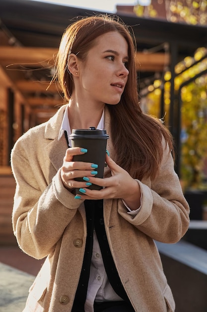 Young caucasian woman in beige coat with long hair and cup of coffee outdoors on sunny day