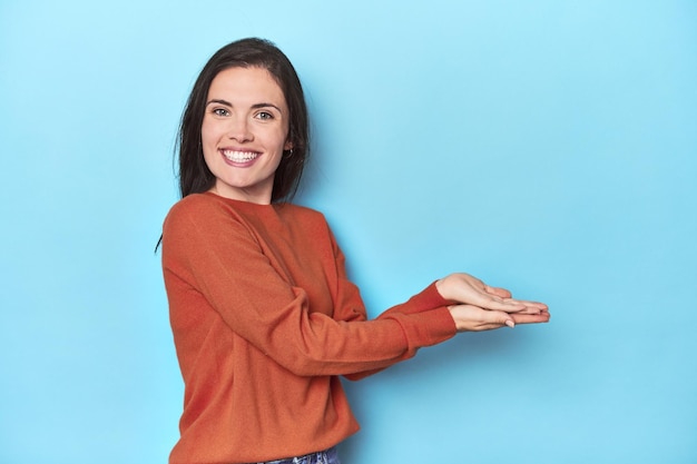 Young caucasian woman on blue backdrop holding a copy space on a palm