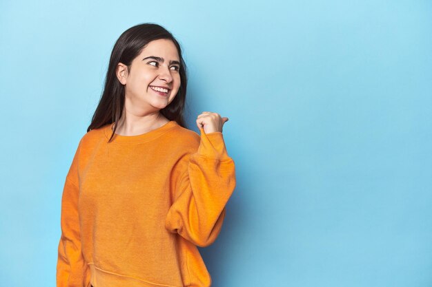 Young Caucasian woman on blue backdrop points with thumb finger away laughing and carefree