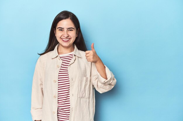 Young Caucasian woman on blue backdrop smiling and raising thumb up