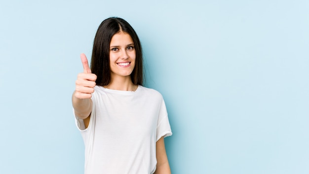 Young caucasian woman on blue wall smiling and raising thumb up