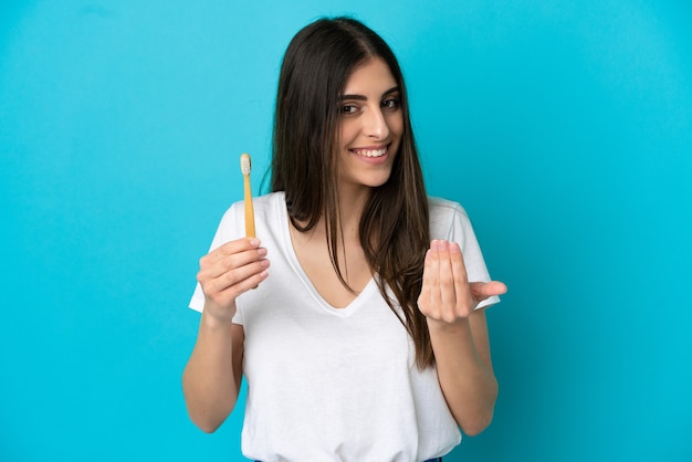 Young caucasian woman brushing teeth isolated on blue background inviting to come with hand. Happy that you came