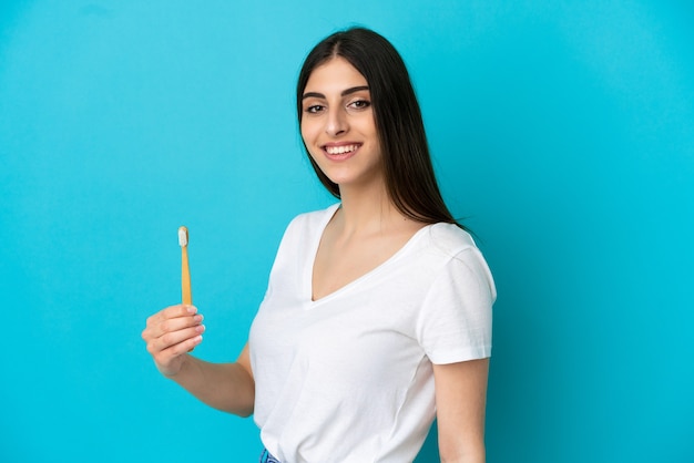 Young caucasian woman brushing teeth isolated on blue background smiling a lot