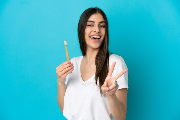 Young caucasian woman brushing teeth isolated on blue background smiling and showing victory sign