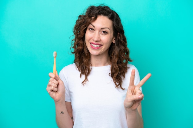 Young caucasian woman brushing teeth isolated on blue background smiling and showing victory sign