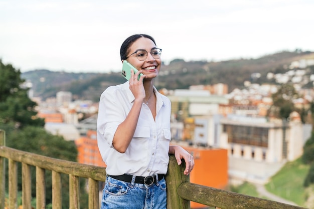 Young Caucasian woman chatting on phone in park with stunning aerial view of city in background under perfect lighting