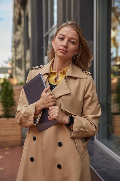 Young caucasian woman in coat holding tablet laptop or ebook in hands outdoors