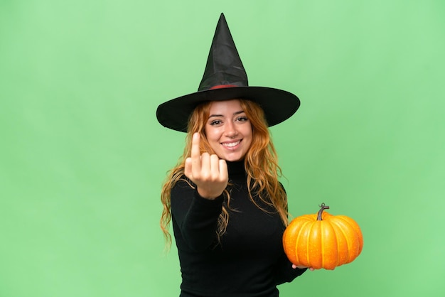 Young caucasian woman costume as witch holding a pumpkin isolated on green screen chroma key background doing coming gesture