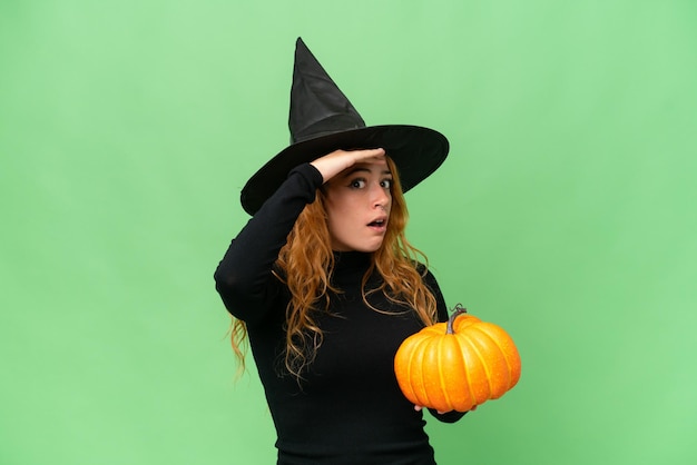 Young caucasian woman costume as witch holding a pumpkin isolated on green screen chroma key background doing surprise gesture while looking to the side