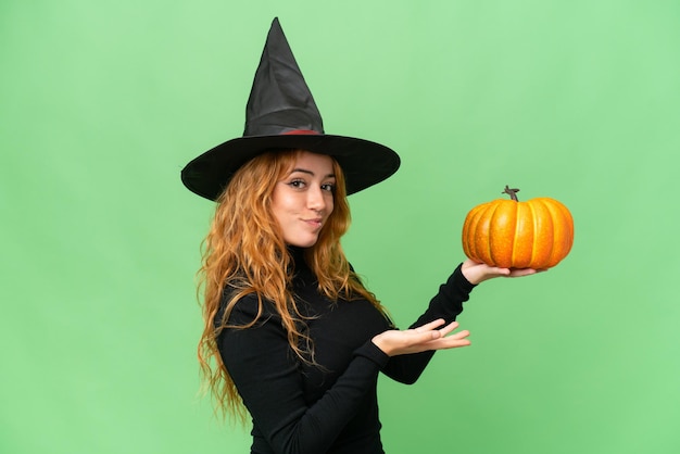 Young caucasian woman costume as witch holding a pumpkin isolated on green screen chroma key background extending hands to the side for inviting to come