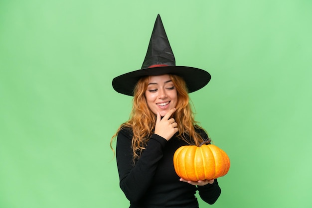 Young caucasian woman costume as witch holding a pumpkin isolated on green screen chroma key background looking to the side and smiling