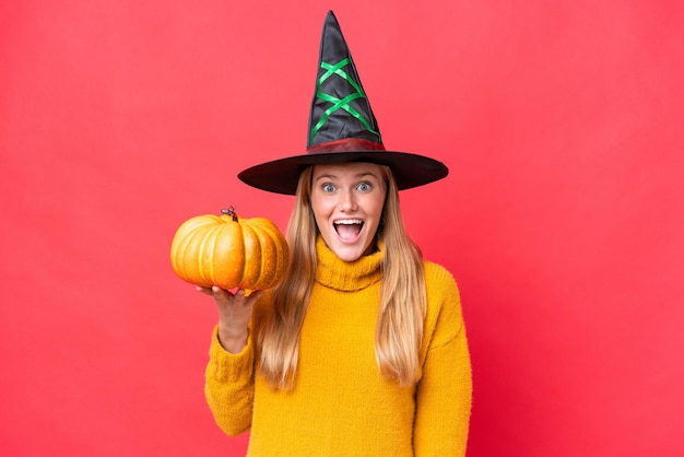 Young caucasian woman costume as witch holding a pumpkin isolated on red background with surprise and shocked facial expression