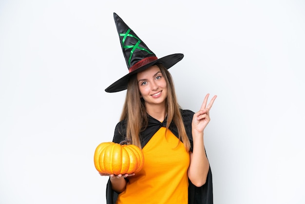 Young caucasian woman costume as witch holding a pumpkin isolated on white background smiling and showing victory sign