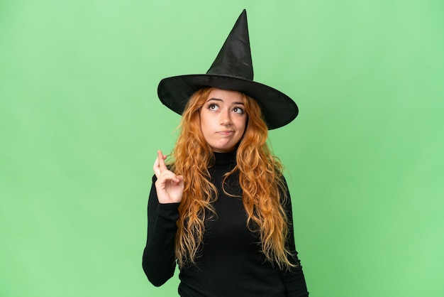 Young caucasian woman costume as witch isolated on green screen chroma key background with fingers crossing and wishing the best