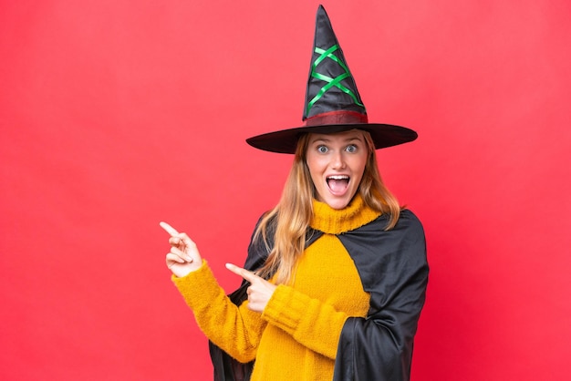 Young caucasian woman costume as witch isolated on red background surprised and pointing side