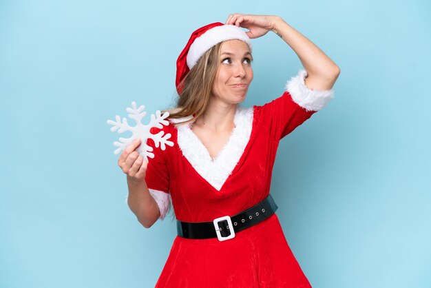 Young caucasian woman dressed as Mama Claus holding a snowflake isolated on blue background having doubts and with confuse face expression