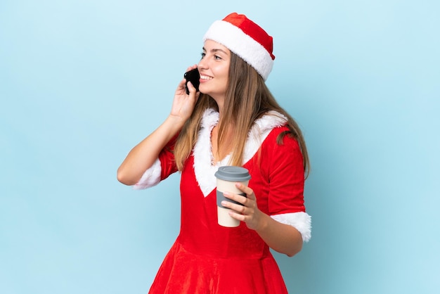 Young caucasian woman dressed as mama noel isolated on blue background holding coffee to take away and a mobile