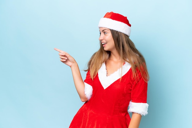 Young caucasian woman dressed as mama noel isolated on blue background pointing finger to the side and presenting a product