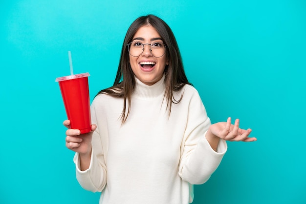 Young caucasian woman drinking soda isolated on blue background with shocked facial expression