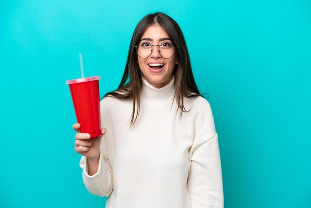 Young caucasian woman drinking soda isolated on blue background with surprise facial expression