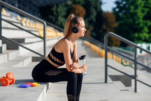 Young caucasian woman drinks water and listen to music after training.