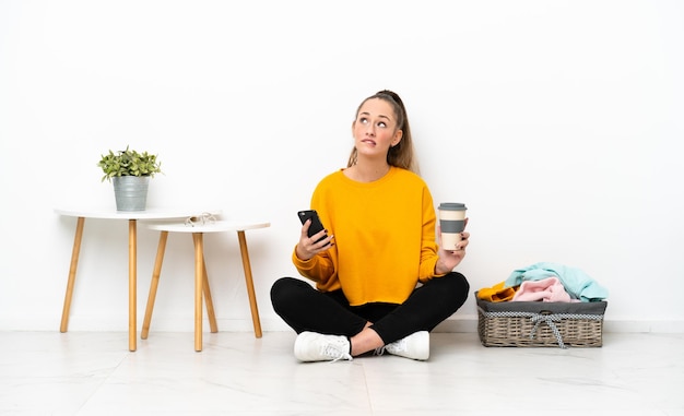 Young caucasian woman folding clothes sitting on the floor isolated on white background holding coffee to take away and a mobile while thinking something