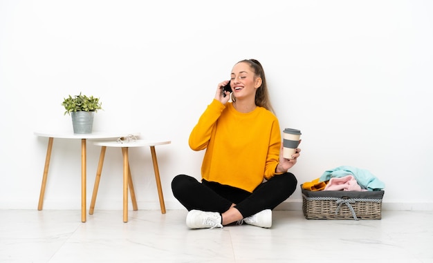 Young caucasian woman folding clothes sitting on the floor isolated on white background holding coffee to take away and a mobile