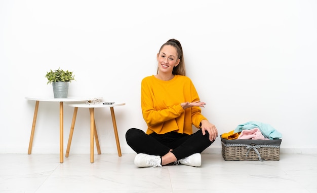 Young caucasian woman folding clothes sitting on the floor isolated on white background presenting an idea while looking smiling towards