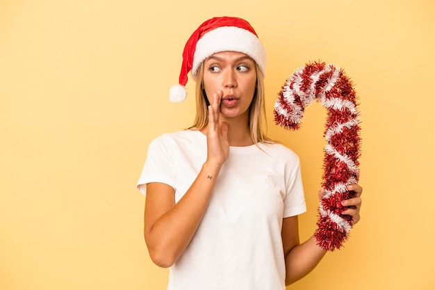 Young caucasian woman holding big christmas stick isolated on yellow background is saying a secret hot braking news and looking aside