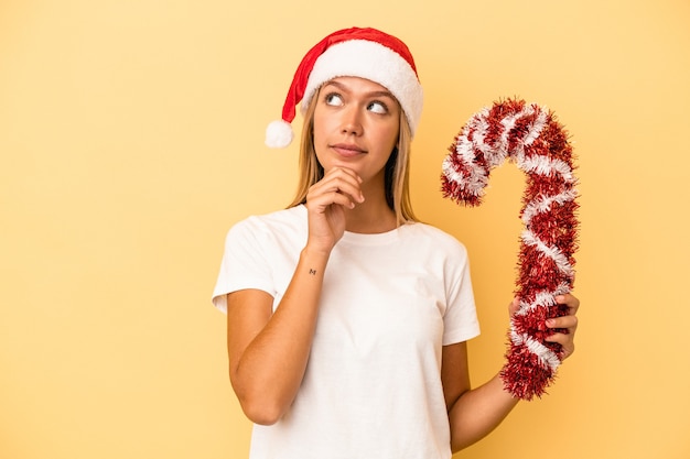 Young caucasian woman holding big christmas stick isolated on yellow background looking sideways with doubtful and skeptical expression.