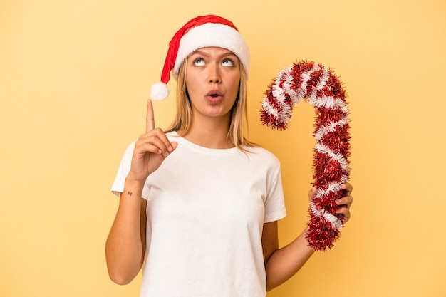 Young caucasian woman holding big christmas stick isolated on yellow background pointing upside with opened mouth.