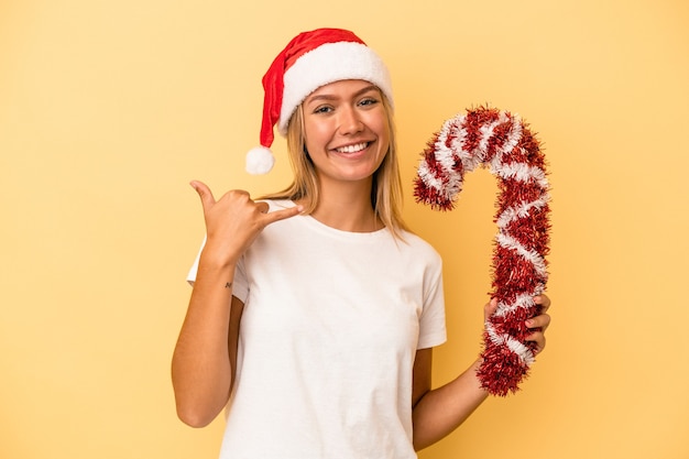 Young caucasian woman holding big christmas stick isolated on yellow background showing a mobile phone call gesture with fingers.