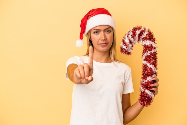 Young caucasian woman holding big christmas stick isolated on yellow background showing number one with finger.