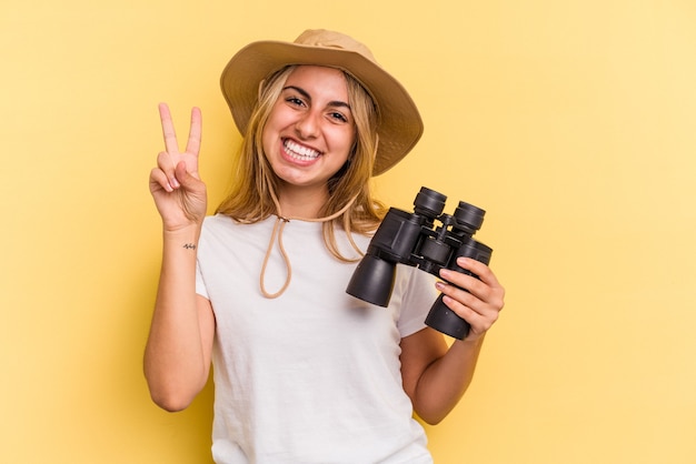 Photo young caucasian woman holding binoculars isolated on yellow background  showing number two with fingers.